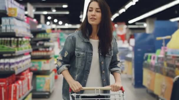 Mulher atraente está dirigindo carrinho de compras através do departamento de alimentos no supermercado e olhando ao redor. Meninas bonitas, vida cotidiana e conceito de compra de produtos . — Vídeo de Stock