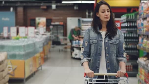 Menina bonita está andando pelo corredor no supermercado com carrinho de compras olhando prateleiras com produtos, os funcionários de uniforme estão trabalhando no fundo . — Vídeo de Stock
