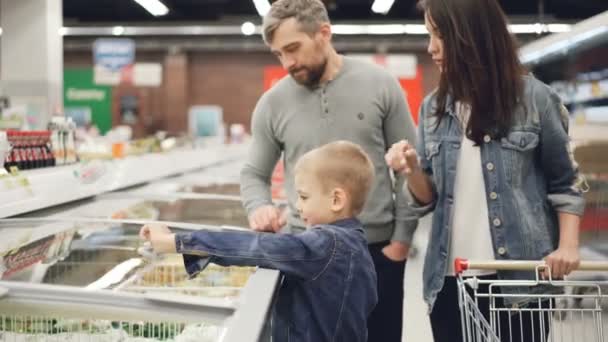 El niño está abriendo el congelador y tomando un paquete de verduras congeladas y luego dándoselo a su papá mientras compra en el supermercado con su familia. Concepto de gente y tienda . — Vídeos de Stock