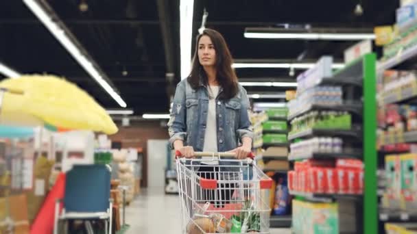 Dolly tiro de menina bonita com carrinho de compras andando ao longo de fileiras de prateleiras com produtos e olhando ao redor com sorriso. Compras de comida e conceito de pessoas . — Vídeo de Stock