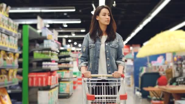 Jeune brune jolie marche dans l'hypermarché et pousser chariot d'achat va acheter de la nourriture et regarder autour des étagères avec des produits. Concept de personnes et commerces . — Video