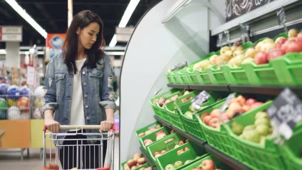 カジュアル衣料に魅力的な女の子はフルーツ行移動歩いてショッピング トロリーと笑顔で有機果実を見てします。健康的なライフ スタイルとスーパー マーケットの概念. — ストック動画
