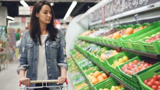Mooie jonge vrouw loopt langs fruit en plantaardige rij duwen winkelwagentje en kijken naar biologisch voedsel met glimlach. Gezonde levensstijl en supermarkt concept. — Stockvideo