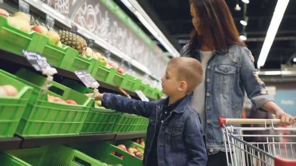 愛らしい小さな男の子が食料品店の青果部門でリンゴを選んでいる、魅力的な母親、彼のそばに立っている、彼女の息子に話して. — ストック動画