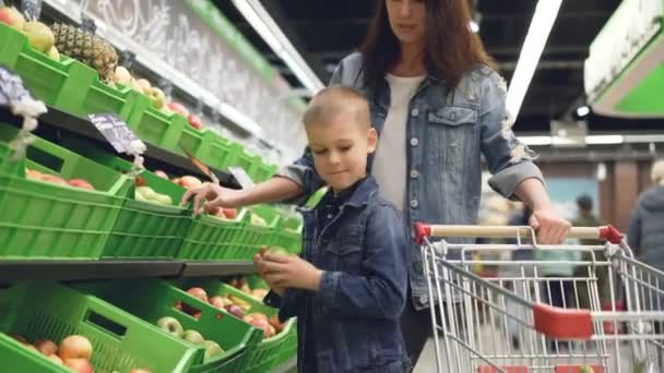 Schattige blonde jongen is winkelen met zijn moeder fruit kopen, hij is apple van plastic doos nemen en brengen het in kar, zijn moeder is lachend en in gesprek met haar zoon. — Stockvideo