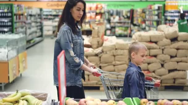 Вид сбоку веселой семейной мамы и сына, гуляющих в супермаркете с торговым троллем, выбирающим фрукты и разговаривающим. Родительство, детство и покупка концепции питания . — стоковое видео
