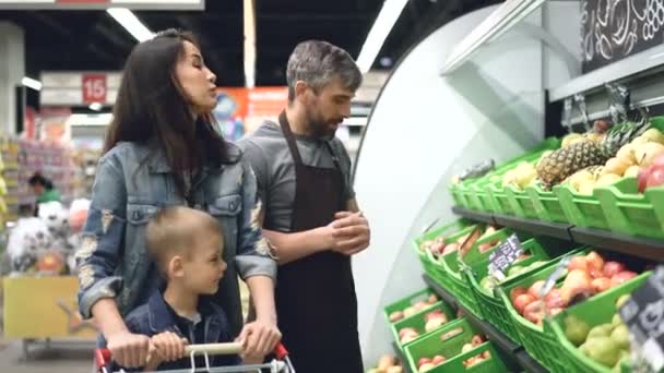 Adjoint de magasin beau sociable vend des fruits frais à la jeune femme attrayante avec enfant, l'homme pointe vers des boîtes lumineuses avec des produits et parlant . — Video