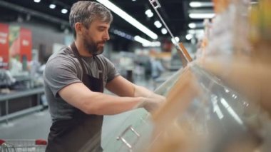 Önlük ciddi satıcı süpermarket gıda kaplarda görüntüleme çalışmaktadır. Rafları ürünleri ile görülebilir, müşteriler yük arabaları ve sepetleri ile dolaşan.