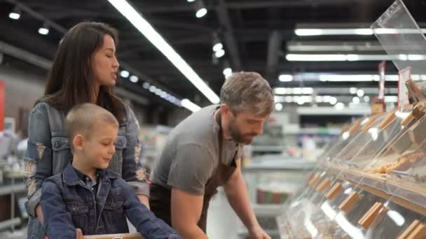 Madre e hijo están comprando panecillos horneados en el supermercado, el vendedor en delantal está poniendo comida en una bolsa de plástico y dándosela a los clientes. Venta y compra de concepto de alimentos . — Vídeo de stock