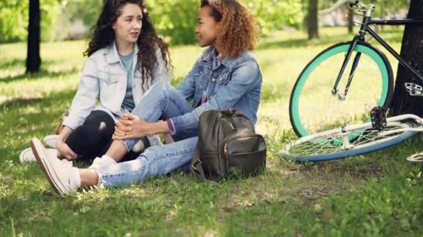 魅力的なアフリカ系アメリカ人の女の子が 公園の芝生の上に座って彼女の白人の友人とチャット友達の話をし笑顔します 現代自転車とバックパックが表示されます — ストック動画