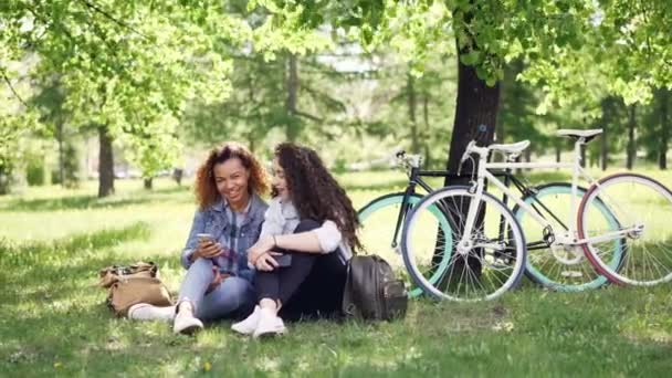 Mulher afro-americana feliz está mostrando seu amigo caucasiano sua tela do smartphone, as meninas estão olhando para ele, falando e rindo. Bicicletas, grama e árvores são visíveis . — Vídeo de Stock