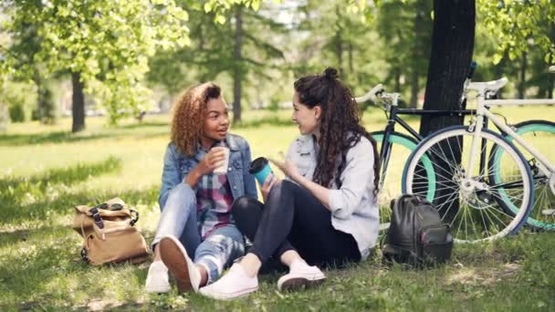 Chicas felices están hablando y bebiendo café para llevar al aire libre sentado en el césped en el parque con mochilas y bicicletas en el fondo. Estilo de vida activo y concepto de personas . — Vídeos de Stock