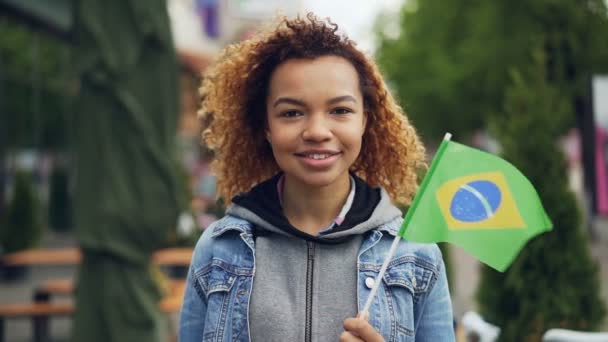 Словоохотливый портрет жизнерадостной афроамериканской девушки, смотрящей в камеру и держащей в руках бразильский флаг, стоящий в красивом парке современного города. Концепция туризма и людей . — стоковое видео