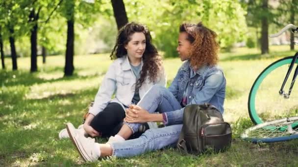 Prachtige vrouwelijke toeristen vrienden in denim kleding ondervindt rest na het paardrijden fietsen, meisjes zitten op gras in park en praten. Fietsen en rugzakken zijn zichtbaar. — Stockvideo