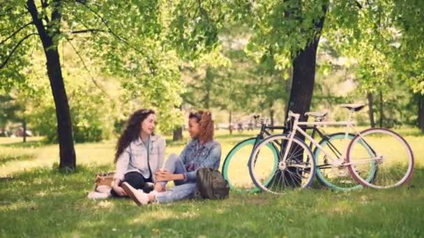 Mosolygó lányok, akiknek beszélgetés park ül a gyep és megosztása Hírek után lovaglás kerékpárok, fiatal nők boldog és nyugodt. Kevert barátság és a természet koncepció. — Stock videók