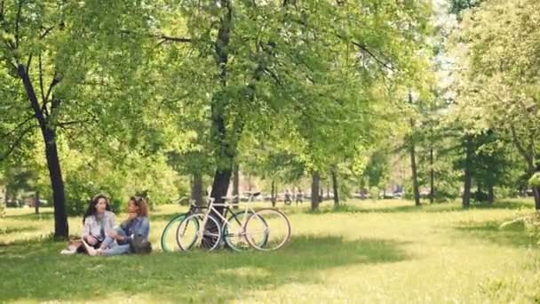 Dos amigas guapas están hablando y riendo sentadas en el césped en el parque en el día soleado de advertencia en verano. Bicicletas modernas y hermosa naturaleza son visibles . — Vídeos de Stock