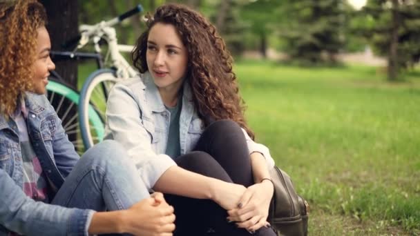 Menina caucasiana está falando com seu amigo afro-americano bonito sentado no parque na grama com bicicletas no fundo. Comunicação, estilo de vida ativo e conceito de pessoas . — Vídeo de Stock