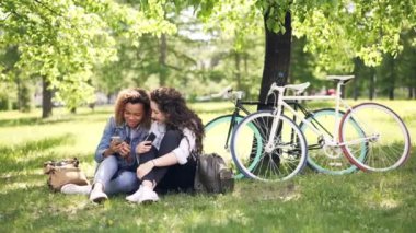 Neşeli genç kadınlar beyaz ve Afrikalı-Amerikalı konuşuyor ve akıllı telefonlar parkta kullanarak, kızlar ekran izlemek ve gülüyor. Bisiklet arka planda vardır.