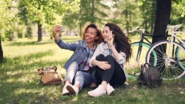 Güzel kızlar arkadaş selfie Park'taki Bisiklet arka planda ile çimenlikte oturan alıyor. Karışık yarış dostluk, modern teknoloji ve neşeli insanlar kavramı.