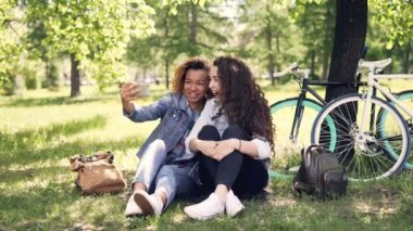 Neşeli kadınlar akıllı telefon ekranına bakarak ve gülüyor sonra selfie birlikte parkta çim üzerinde oturan kamera için poz alarak. Bisiklet ve sırt çantaları görülebilir.