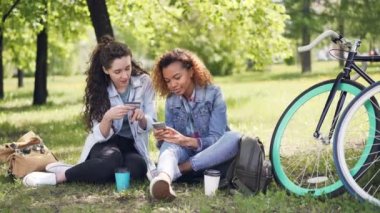 İki genç kadın kart kredi kartı tutan çimenlerin üzerinde parkta oturan smartphone kullanarak ödeme ve telefon o zaman çak bir beşlik yapıyor ve gülüyor. Online ödeme kavramı.