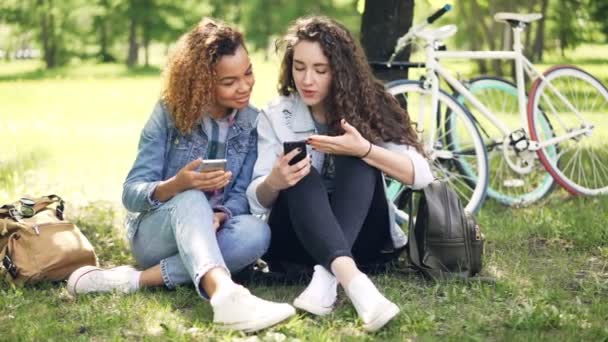 Привабливі дівчата в джинсовому одязі використовують смартфони, дивлячись на екран, розмовляючи і сміючись, сидячи в парку на траві. Видимі велосипеди та рюкзаки . — стокове відео