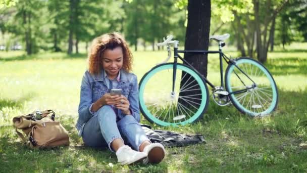 Gelukkig African American girl is het gebruik van smartphone scherm aan te raken en glimlachen terwijl rust in park na rijden fiets. Moderne tas en fiets zijn zichtbaar. — Stockvideo