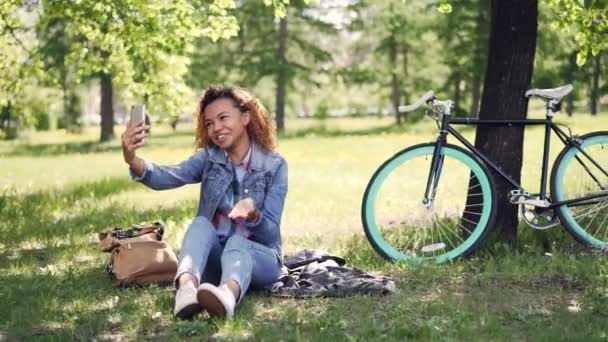 Menina muito afro-americana está fazendo videochamada com smartphone, ela está olhando para a tela, falando e acenando a mão. Belo parque com grama e árvores é visível . — Vídeo de Stock