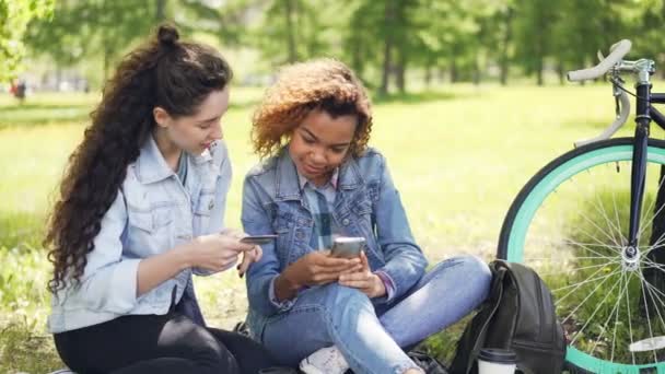 Joven mujer afroamericana está de compras en línea haciendo el pago con el teléfono inteligente, mientras que su amigo en la celebración de la tarjeta de crédito, las niñas están sentadas en el césped en el parque en verano . — Vídeo de stock