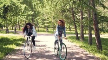 Afro-Amerikan kız beyaz arkadaşıyla beraber şehir parkı içinde bisiklete biniyor, Genç kadınlar konuşmaya ve birlikte gülüyor. Güzel doğa, güneşli bahar günü.