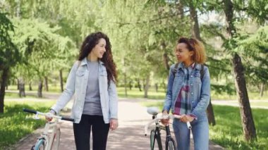 İki bisikletçiler Afrikalı-Amerikalı ve beyaz bisiklet güzel yeşil parkın içinden ile yürüme, konuşuyor, gülüyor ve yaz ve güneşli bir gün zevk.
