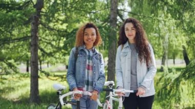 Mutlu genç kadınlar beyaz ve güzel yeşil parkta birlikte ayakta, Bisiklet holding, kameraya bakıyor ve gülümsüyor Amerikan Afrika portresi. İnsanlar ve doğa kavramı.