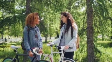 İki güzel kadın bicyclers sırt çantaları ile ayakta tutan Bisiklet Park konuşuyor. Güzel şehir parkı birçok ağaç ve yeşil çimenler kadeh kaydırmak.