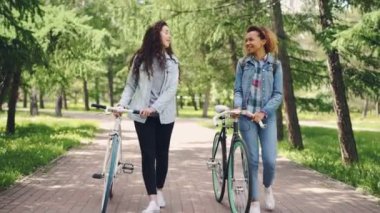 Sportif genç kadın Bisiklet ile parkta yürüyüş ve zevk güzel çam ağaçları ve ılık bahar günü sohbet. Onlar trendy denim giysiler giyiyorsun.