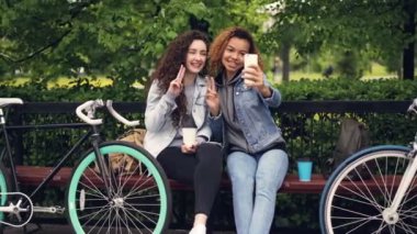 Çok dikkatsiz kızlar selfie poz ve bisiklet ile parkta oturan gitmek kahve tutan smartphone ile alıyor. Modern yaşam tarzı, dostluk, mutlu insanlar ve teknoloji kavramı.
