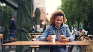 Mutlu Afro-Amerikan kız ekran dokunaklı ve açık park kafede otururken gülümseyen smartphone kullanıyor. Sırt çantası ve bisiklet görülebilir.