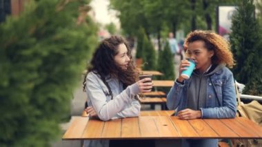 Çekici Kafkas ve Afrikalı-Amerikalı kız arkadaş kahve içme ve şehir parkı güzel ağaçlar ile açık kafede oturan sohbet.