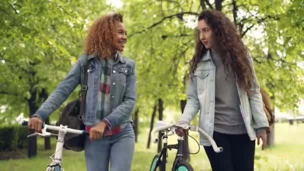 Щасливі друзі красиві жінки гуляють в парку, тримаючи велосипеди і весело розмовляючи, красиві природні дерева і трава на задньому плані. Концепція дружби та хобі . — стокове відео