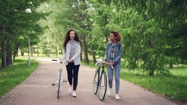 Dolly disparo de hermosas niñas afroamericanas y caucásicas caminando en el parque a lo largo del camino con bicicletas y hablando. Árboles verdes y césped son visibles . — Vídeo de stock