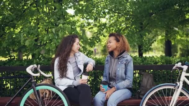 Atractivas mujeres jóvenes caucásicas y afroamericanas están charlando y bebiendo café para llevar mientras están sentadas en el banco en el parque y descansando después de montar en bicicleta . — Vídeo de stock