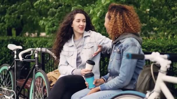 Znajomych szczęśliwy dziewcząt są rozmawiając i śmiejąc się, trzymając kawy na wynos, siedząc na ławce w parku z rowerami. Nowoczesny styl życia, aktywnego życia miejskiego, koncepcja wesoły osób. — Wideo stockowe