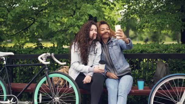 Cheerful Caucasian and African American girls friends are taking selfie making moustache from hair and posing with finger gestures sitting on bench in park. — Stock Video