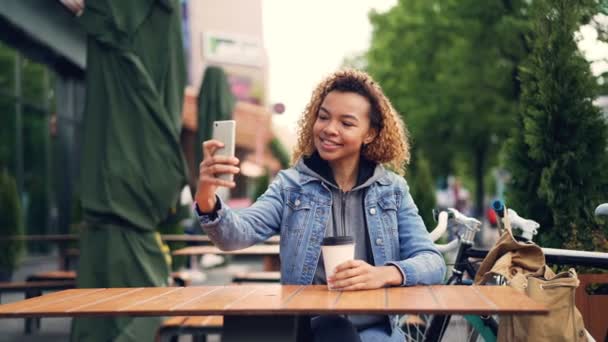 Nieostrożny African American kobieta zadowolony turysta jest biorąc selfie z smartphone w kawiarni na świeżym powietrzu siedzi przy stole z kawy na wynos, rower i plecak są widoczne. — Wideo stockowe