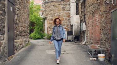 Afro-Amerikan kız memnun turist sırt çantası ile yürüyüş ve kameraya bakarak taş duvarlar ile sokakta dans mutlu. İnsanlar ve Turizm kavramı.