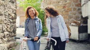 Slowmotion gülüyor ve eski binalar ile cadde boyunca bisiklet ile yürüyüş mutlu Kafkasya ve Afrikalı-Amerikalı kızlar turist. Turizm ve insanlar kavramı.