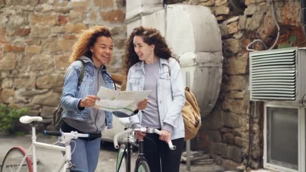 Aufgeregte junge Fahrradfahrerinnen schauen auf die Karte und schauen sich lachend um, während sie mit Fahrrädern auf der Straße in der Fremde stehen. Menschen und Reisekonzept. — Stockvideo