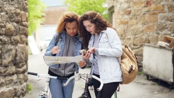 Verlorene Touristen Hübsche Mädchen schauen auf Papierkarte und Smartphone-Bildschirm, die mit Fahrrädern auf der Straße stehen und nach einem Touristenziel suchen. — Stockvideo
