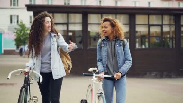 Dolly shot of attractive girls friends walking with bicycles along street with beautiful old buildings and talking happily. Active lifestyle and tourism concept. — Stock Video