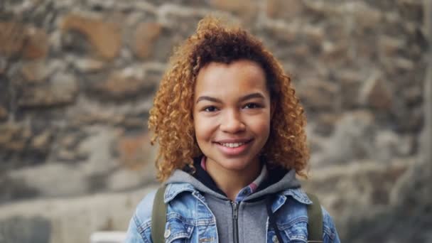 Retrato de perto da menina afro-americana com cabelo encaracolado claro vestindo roupas casuais olhando para a câmera e sorrindo. Jovens felizes e conceito de estilo de vida moderno . — Vídeo de Stock