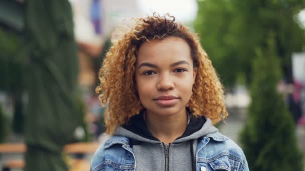 Slowmotion porträtt av ganska African American girl moderna tonåring kamera och tittar och ler medan stående i vacker grön park. Glada människor och koncept. — Stockvideo